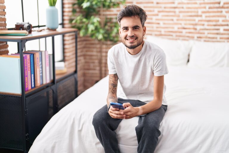 young-hispanic-man-using-smartphone-sitting-bed-bedroom-min