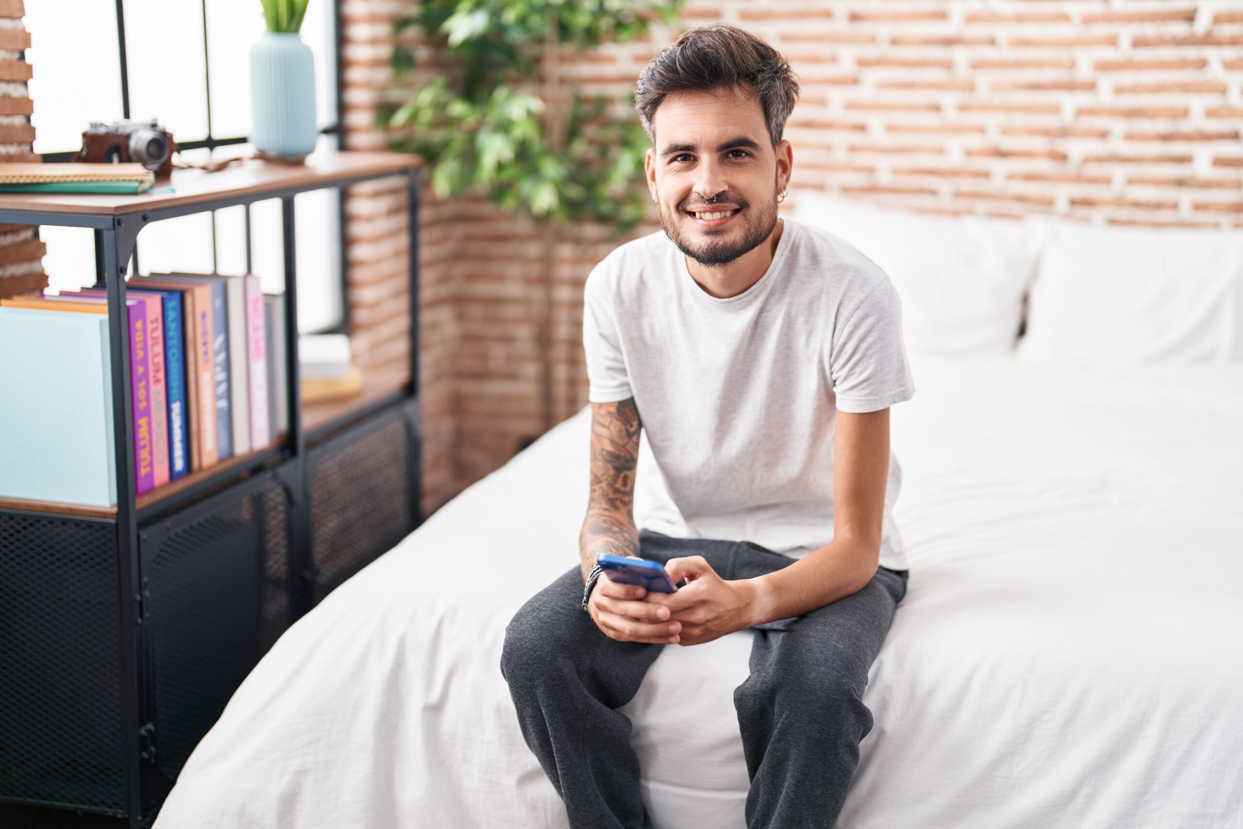 young-hispanic-man-using-smartphone-sitting-bed-bedroom-min